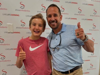 Boy smiling showing off his braces with an adult next to him with both of their thumbs up